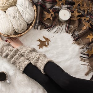Op de afbeelding: Een paar bruine gebreide beenwarmers met zwarte leggings op een wit pluche oppervlak. Op de achtergrond een mand met garenbollen en een brandende kaars.