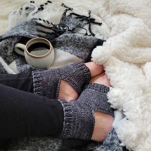 Puede incluir: Un par de guantes sin dedos de punto gris con un agujero para el pulgar, usados por una persona sentada en una manta blanca y negra con una taza de café y gafas.