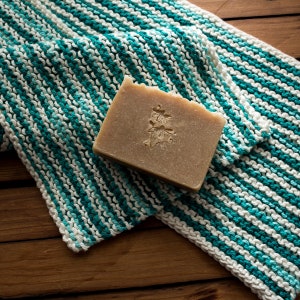 May include: A bar of natural soap with a plant design on a white and teal striped knitted washcloth.