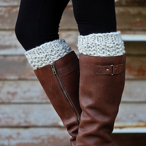 May include: A pair of brown leather boots with a zipper and buckle detail. The boots are worn with black leggings and cream-colored knit leg warmers. The leg warmers have a textured, chunky knit design.