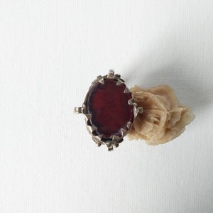 Può includere: Un anello d'argento con una pietra ovale rosso scuro. L'anello ha un bordo decorativo e frastagliato attorno alla pietra. Un oggetto beige a forma di rosa è accanto all'anello su una superficie bianca. Gioielli.