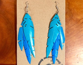 May include: A pair of blue feather earrings with silver hooks. The earrings are made from recycled materials and are part of a collection that promotes sustainability. The tag reads "rethink recycle reuse".