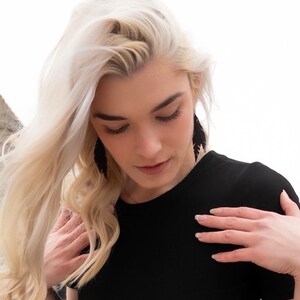 May include: A woman with long, blonde hair wearing a black, short-sleeved shirt and black feather earrings. The woman is looking down at her shirt.