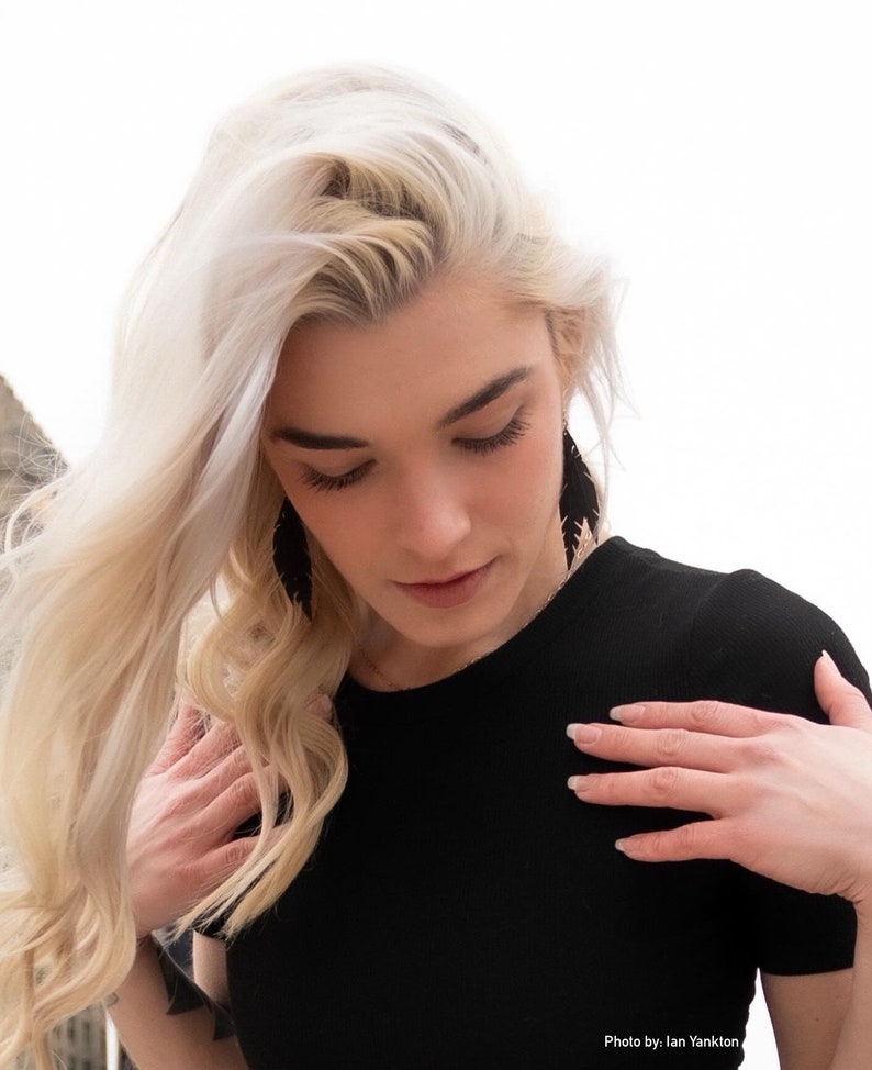 May include: A woman with long, blonde hair wearing a black, short-sleeved shirt and black feather earrings. The woman is looking down at her shirt.