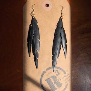 May include: A pair of black feather earrings made from recycled tire inner tubes. The earrings are hanging from gold hooks and are displayed on a brown paper tag with the text "rethink recycle reuse" and a logo with a stylized "R" and a number "3" inside a circle.