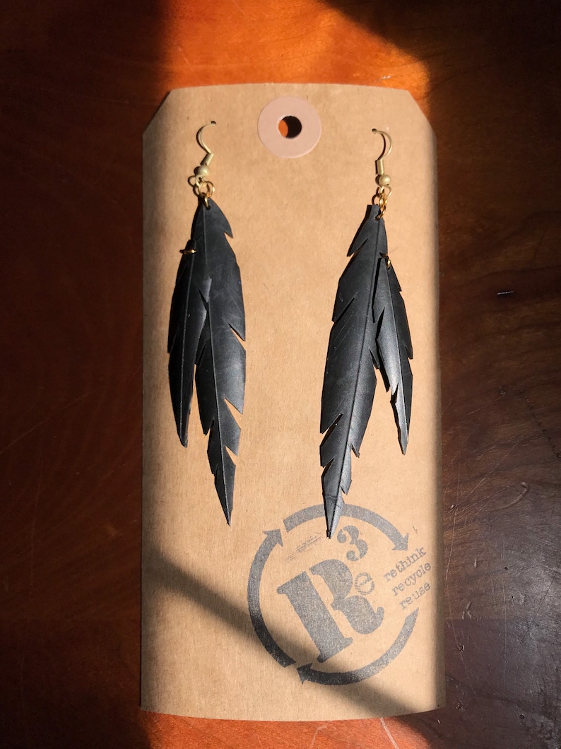May include: A pair of black feather earrings made from recycled tire inner tubes. The earrings are hanging from gold hooks and are displayed on a brown paper tag with the text "rethink recycle reuse" and a logo with a stylized "R" and a number "3" inside a circle.