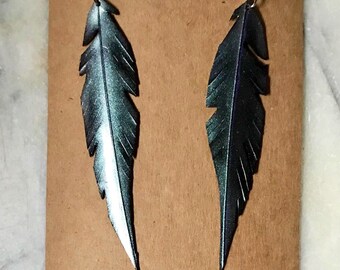May include: A pair of dangle earrings featuring a feather design made from recycled materials. The feathers are a dark green and silver color with a metallic sheen. The earrings are hanging from a brown card with the text "rethink recycle reuse."