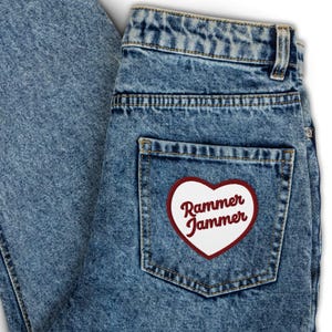 Könnte beinhalten: Nahaufnahme einer hellblauen Jeans. Die Gesäßtasche hat einen herzförmigen Aufnäher mit weißem Zentrum, rotem Rand und dem Schriftzug "Rammer Jammer" in Schreibschrift.