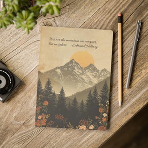 May include: A notebook with a mountain landscape design, featuring a quote by Edmund Hillary. The cover art includes a mountain range, a setting sun, and a forest. A pencil and pen are placed next to the notebook on a wooden surface.