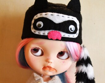 May include: A doll with long pink and blue hair wearing a black and white raccoon hat with a pink nose. The doll is wearing a beige sweater and striped pants.