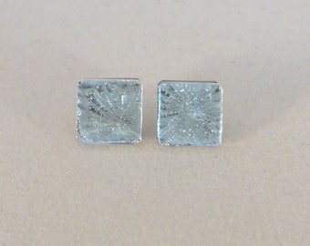 May include: A pair of square, pale blue stud earrings. Each earring has a textured, crystalline appearance. The earrings are presented against a neutral backdrop, emphasising their unique design. These earrings are a simple yet elegant accessory.