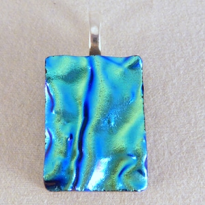 May include: Rectangular dichroic glass pendant with a silver bail. The pendant features a vibrant, iridescent surface with shades of blue and green. The glass has a textured, rippled appearance, creating a unique visual effect. The bail is a simple, rectangular loop.