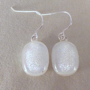 May include: A pair of elegant, oval-shaped earrings. Each earring features a clear, iridescent, glitter-filled oval pendant suspended from a silver-colored hook. The earrings have a simple, yet sophisticated design.