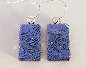 May include: A pair of rectangular earrings with a blue and green iridescent pattern. The earrings have a silver-colored metal frame and ear wires. The design features a textured, fan-like pattern within the blue glass.