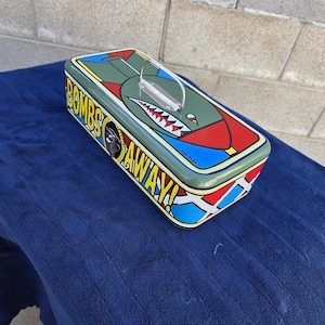 May include: A rectangular tin box with a military theme. The box is olive green with a red and blue design on the ends. The top features a shark mouth design and the words "BOMBS AWAY!" in yellow.