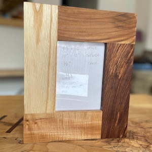 May include: A multi-toned wooden picture frame. The frame is made of walnut, butternut, silver maple, and ash. Inside dimensions are 11.4 cm x 16.5 cm, outside dimensions are 25.4 cm x 30.5 cm, and the thickness is 1.9 cm.