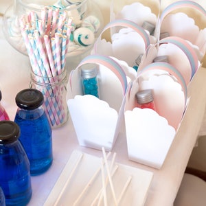 Könnte beinhalten: Eine Tischdekoration für eine Party mit Regenbogen-Thema. Es gibt weiße Geschenkboxen mit Regenbogen-Besatz, blaue und rosa Süßigkeiten in kleinen Glasflaschen, blaue und rosa Getränke in Glasflaschen und Marshmallow-Lollies mit Streuseln auf einem weißen Teller.