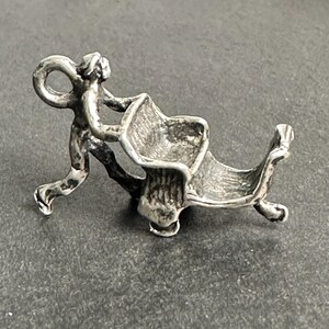 May include: A silver-toned metal charm depicting a figure pushing a wheelbarrow. The figure is shown in a walking stance with arms extended forward, gripping the handles of the wheelbarrow, which features a deep basin and a single front wheel.