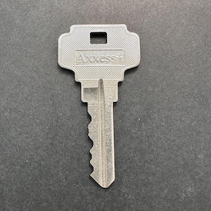 May include: A silver-colored key with the text "Axxess+" on the head. The key has a rectangular head with a small square hole and a ridged pattern. The key's blade has a unique, wavy edge.