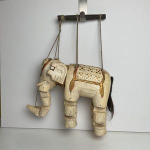 May include: A vintage, off-white wooden elephant puppet with articulated joints. The puppet has gold and red accents, a black tail, and is suspended by strings from a wooden bar. The elephant's trunk is extended, and it has a decorative saddle.