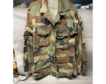 May include: Camouflage pattern military jacket with multiple pockets and the text "O'DOWD" and "U.S. ARMY" on the front. The jacket features a button-up front and a collar. The color palette includes shades of green, brown, and black.