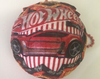 May include: A red, white, and black fabric ornament with a Hot Wheels logo and a red race car design. The ornament has a gold trim and a beaded chain.