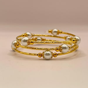 May include: Gold-toned spiral bracelets with white faux pearls. The bracelets have a textured design and are adorned with small gold beads. The jewelry is displayed on a light pink surface.
