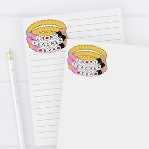 Puede incluir: Dos blocs de notas blancos con papel rayado. El bloc de notas superior tiene una pila de pulseras de cuentas de colores con las palabras "Me encanta mi &eacute;poca de maestra" en ellas. El bloc de notas inferior tiene las mismas pulseras y el texto "desde el escritorio de la Sra. ANDERSON".