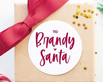 May include: A wrapped gift box with a red ribbon and a white circular sticker that reads "To Brandy Santa" in a cursive font. Gold star-shaped confetti is scattered around the box, suggesting a festive occasion.