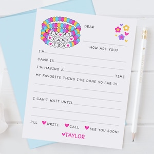May include: A white card with a pink and blue bracelet charm with the words "In My Camp Era" on it. The card has a fill-in-the-blank letter format with the text "Dear", "How are you?", "I'm", "Camp is", "I'm having a", "My favorite thing I've done so far is", "I can't wait until", "I'll", "Write", "Call", "See you soon!" and "♥Taylor".