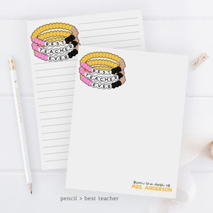 Puede incluir: Una tarjeta blanca con una pulsera de cuentas rosa, negra y dorada que dice "Mejor Maestro de Todos los Tiempos". La tarjeta tambi&eacute;n tiene el texto "desde el escritorio de MRS. ANDERSON" y "l&aacute;piz > mejor maestro" en la parte inferior.