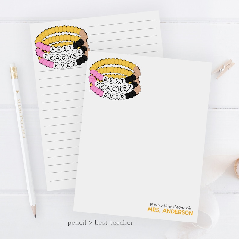 Puede incluir: Una tarjeta blanca con una pulsera de cuentas rosa, negra y dorada que dice "Mejor Maestro de Todos los Tiempos". La tarjeta tambi&eacute;n tiene el texto "desde el escritorio de MRS. ANDERSON" y "l&aacute;piz > mejor maestro" en la parte inferior.