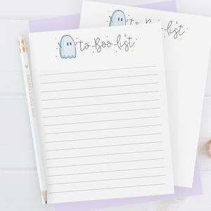 May include: A stack of lined paper with a ghost illustration and the words "to-Boo-list" in a whimsical font. A white pencil with gold accents and the text "Pink Pickle Parties" is on the left. The paper is on a light purple background.