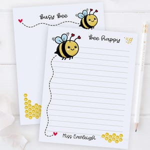 May include: Two white notepads with bee-themed illustrations. One notepad says "busy bee" with a cartoon bee and a dotted line. The other says "bee happy" with a bee, lined paper, and the name "Miss Everleigh". A pencil is on the right.