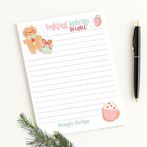 May include: A lined notepad with the text "baking spirits bright" and illustrations of a gingerbread man, a gift, a jar of jam, and a cup of hot cocoa. A black pen and a sprig of greenery are on the white surface.