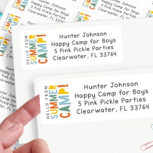 May include: White rectangular address labels with the text "HELLO FROM SUMMER CAMP!" in orange, blue, and yellow. The labels include the name "Hunter Johnson" and camp details. The image shows a hand touching the labels.