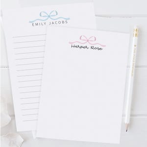 May include: Two white notepads with bow designs. One notepad has a blue bow and the name "Emily Jacobs" printed below. The other notepad has a pink bow and the name "Harper Rose" printed below. A white pencil with gold accents is also present.