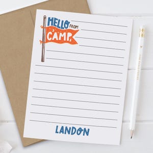 May include: A lined notepad with the text "Hello From Camp" in blue and orange, and the name "Landon" in blue. A white pencil with gold accents and the text "Pine Pickle Parties" is on the right. The notepad rests on a brown envelope.