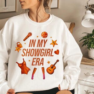 K&ouml;nnte beinhalten: Wei&szlig;es Sweatshirt mit dem Text "IN MY SHOWGIRL ERA" in orangefarbenem Glitzer. Das Design umfasst Illustrationen eines Mikrofons, eines Kleides, einer Gitarre und Sterne. Ein Herz und ein Schloss sind ebenfalls enthalten.