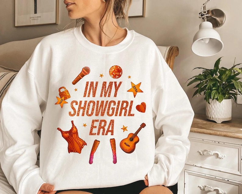 K&ouml;nnte beinhalten: Wei&szlig;es Sweatshirt mit dem Text "IN MY SHOWGIRL ERA" in orangefarbenem Glitzer. Das Design umfasst Illustrationen eines Mikrofons, eines Kleides, einer Gitarre und Sterne. Ein Herz und ein Schloss sind ebenfalls enthalten.