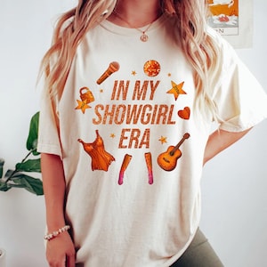 K&ouml;nnte beinhalten: Cremefarbenes T-Shirt mit dem Text "IN MY SHOWGIRL ERA" in Orange. Das Design umfasst orangefarbene Illustrationen eines Mikrofons, einer Discokugel, Sterne, eines Herzens, eines Kleides und einer Gitarre.