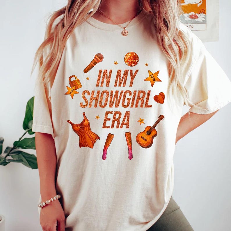 K&ouml;nnte beinhalten: Cremefarbenes T-Shirt mit dem Text "IN MY SHOWGIRL ERA" in Orange. Das Design umfasst orangefarbene Illustrationen eines Mikrofons, einer Discokugel, Sterne, eines Herzens, eines Kleides und einer Gitarre.