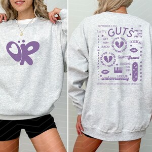 Puede incluir: Una sudadera gris claro con un gran gr&aacute;fico morado "OR" en la parte delantera. La parte trasera presenta texto morado, incluyendo "Lacy Guts" y otras frases. La sudadera es de estilo casual.