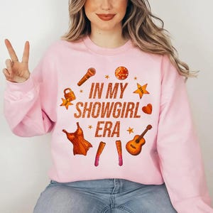 K&ouml;nnte beinhalten: Hellrosa Sweatshirt mit dem Text "IN MY SHOWGIRL ERA" in orangefarbenem Glitzer. Das Design umfasst Illustrationen eines Mikrofons, eines Kleides, einer Gitarre und Sterne. Das Sweatshirt hat eine hellrosa Farbe.