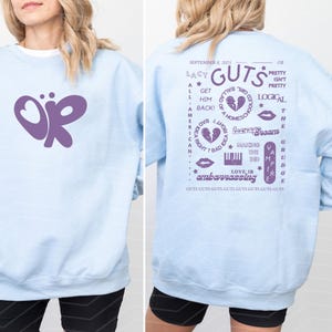 Puede incluir: Sudadera azul claro con un dise&ntilde;o de mariposa morada en la parte delantera. La parte trasera de la sudadera tiene texto morado, incluyendo "Lacy Guts" y otras frases. El dise&ntilde;o es informal.