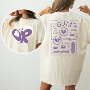 Puede incluir: Camiseta color crema con un gran dise&ntilde;o de mariposa morada y las letras "OR". La parte trasera de la camiseta presenta la palabra "GUTS" y otros textos y gr&aacute;ficos en morado, incluidas las palabras "Lacy", "Pretty" y "Embarrassing".