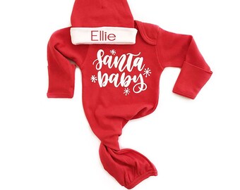 May include: Red baby gown with long sleeves and a white 'Santa Baby' design. The gown is tied at the bottom and comes with a matching red hat with a white band that says 'Ellie'.