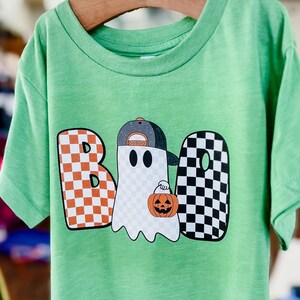 May include: Green short-sleeved t-shirt with a checkered "BOO" graphic featuring a ghost wearing a baseball cap and holding a pumpkin.