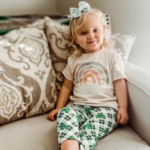 Op de afbeelding: Een jong kind draagt een lichtbeige t-shirt met een regenboog-print en een groen-wit geruite broek met klavertjesvier-ontwerpen. Het kind zit op een bank met een lichtbruin, gepatroneerd kussen.