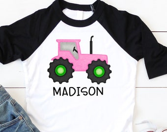 Puede incluir: Una camiseta de béisbol blanca y negra con un diseño de tractor rosa y el nombre "MADISON" impreso en la parte delantera. La camiseta se combina con jeans azules y zapatillas blancas.